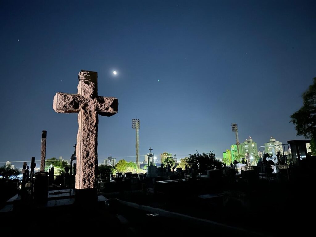Na imagem vemos um cemitério à noite, com uma grande cruz de pedra em primeiro plano, iluminada lateralmente de modo a realçar sua textura rugosa. Primeiro plano: cruz de pedra maciça, posicionada à esquerda, com detalhes e sombras que mostram sua superfície irregular. Ao redor há outras lápides e monumentos mais baixos, em silhueta. Céu e iluminação: céu noturno limpo, em tom azul-escuro; a Lua em fase crescente/brilhante aparece pouco à esquerda do centro superior, e algumas estrelas tênues pontilham o céu. A iluminação artificial (provavelmente de postes ou da cidade) ilumina partes das lápides e da vegetação. Plano de fundo urbano: ao longe há prédios iluminados — alguns com luzes verdes — e postes altos de iluminação (provavelmente de um estádio ou campo), que recortam o horizonte. Árvore escura aparece à direita, enquadrando a cena. Atmosfera: imagem transmite uma sensação tranquila e levemente melancólica, com contraste entre a calma do cemitério e as luzes vivas da cidade ao fundo.