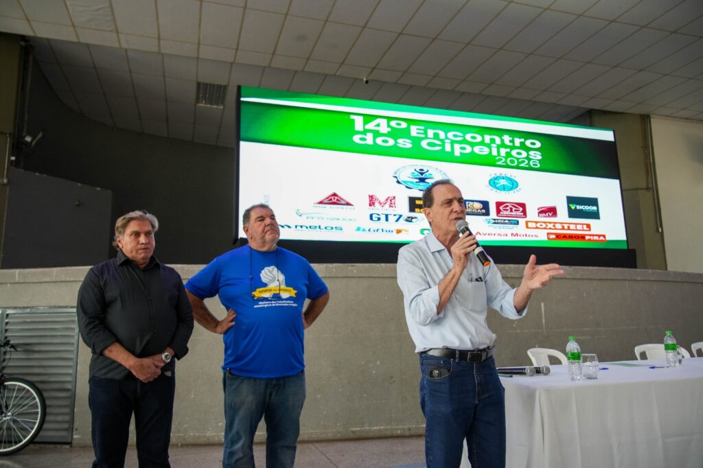 Esta fotografia documenta um momento formal de apresentação durante o 14º Encontro dos Cipeiros em 2026, onde um palestrante discursa em um palco diante de uma plateia ou mesa diretora. A cena é composta de forma centralizada em um palco amplo, destacando três homens posicionados em frente a um grande painel digital. O orador principal encontra-se à direita, segurando um microfone e gesticulando com a mão esquerda, enquanto os outros dois homens permanecem ao lado, ouvindo atentamente. A composição é clássica de eventos corporativos, utilizando o telão como pano de fundo informativo para contextualizar o evento, com uma mesa coberta por uma toalha branca no canto inferior direito, sugerindo uma estrutura de conferência ou reunião de trabalho. Os três homens são adultos, aparentando estar na faixa da meia-idade ou maturidade. O orador, posicionado à direita, veste uma camisa social de cor clara com mangas dobradas e jeans, mantendo uma postura engajada e expressiva. O homem ao centro, com uma postura mais relaxada, veste uma camiseta azul com um logotipo estampado e calças jeans. À esquerda, o terceiro homem utiliza uma camisa preta e calças escuras, mantendo uma postura mais reservada com as mãos juntas à frente. Todos apresentam vestimentas informais de negócios, condizentes com um ambiente de encontro profissional de segurança do trabalho. A imagem é uma fotografia digital capturada com foco nítido e iluminação ambiente clara. A qualidade da imagem reflete uma captura direta de um evento, com cores naturais e uma profundidade de campo que mantém tanto os sujeitos quanto o texto informativo do painel digital em foco. Não há manipulação artística óbvia, tratando-se de um registro documental que prioriza a clareza informativa e a captura precisa do momento da oratória. O cenário é um auditório ou espaço para eventos, caracterizado por paredes de concreto aparente e um palco elevado. A iluminação é composta por luzes de teto distribuídas uniformemente, que iluminam bem os sujeitos e o grande painel digital, o qual exibe o título "14º Encontro dos Cipeiros 2026" acompanhado por diversos logotipos de empresas parceiras em um fundo verde vibrante. O ambiente transmite uma atmosfera de profissionalismo, colaboração e foco, típica de seminários corporativos ou encontros de profissionais de segurança, com a mesa branca adicionando um elemento de formalidade institucional ao espaço.