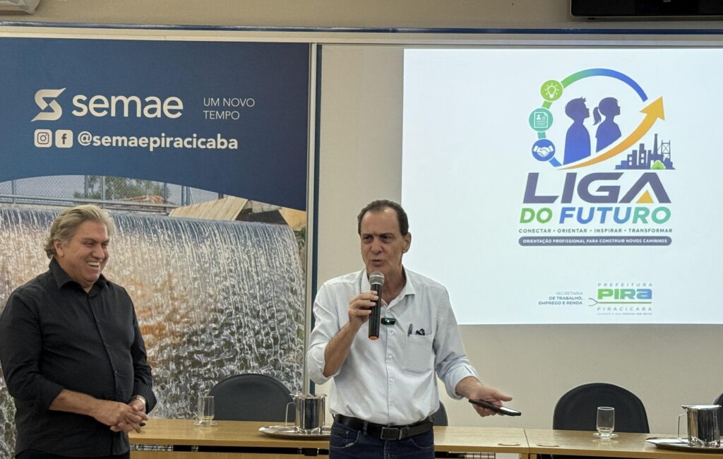 A imagem mostra dois homens em um ambiente de apresentação institucional. Um deles fala ao microfone, enquanto o outro observa sorrindo. Ao fundo, há um telão com o logotipo do projeto “Liga do Futuro”, iniciativa da Prefeitura de Piracicaba voltada à orientação profissional e desenvolvimento de carreira. Também aparecem banners do SEMAE Piracicaba, reforçando a parceria no evento.