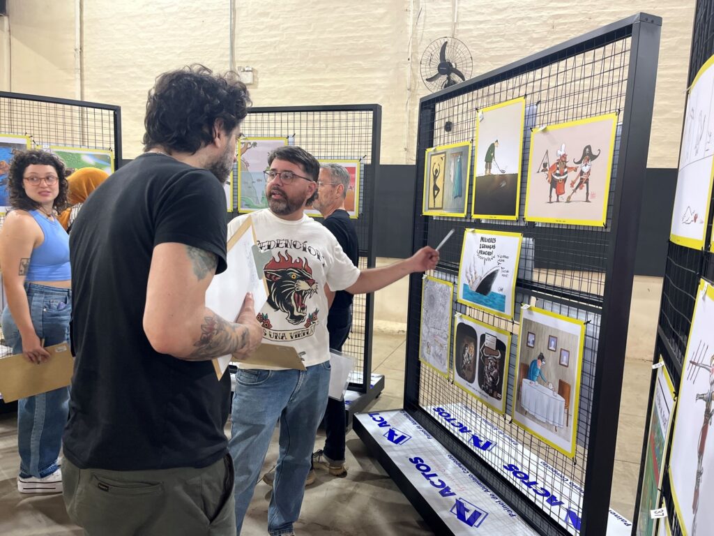 A imagem mostra visitantes em uma exposição de ilustrações, montada em painéis de grades metálicas. No centro, um homem de óculos e barba, usando uma camiseta branca com a estampa de um tigre, aponta para uma das obras enquanto conversa com um homem de camiseta preta, que segura alguns quadros. À esquerda, uma mulher de óculos e blusa azul observa a cena, acompanhada por outra pessoa usando um lenço amarelo.