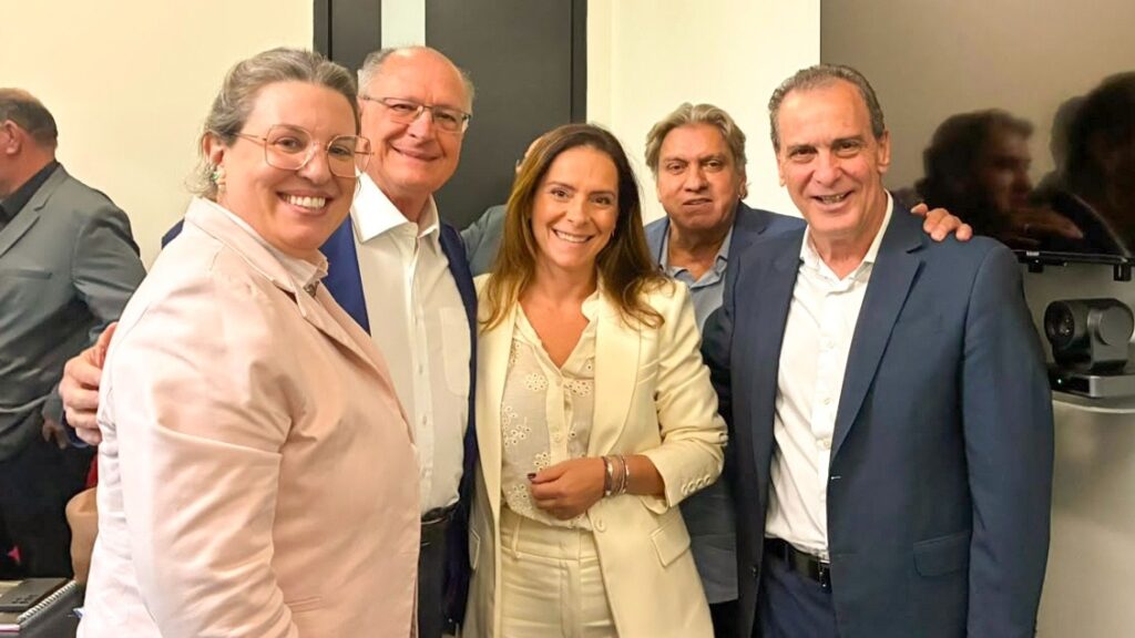 Esta imagem mostra um grupo de cinco pessoas sorridentes, posando para uma foto em um ambiente interno. Aqui estão os detalhes: As Pessoas: Na esquerda, há uma mulher com cabelos presos, usando óculos de armação clara e um blazer rosa claro. Ela sorri abertamente. Ao lado dela, um homem de meia-idade, com cabelos grisalhos e óculos, veste uma camisa branca e um terno azul. Ele também sorri. No centro, uma mulher de cabelos castanhos longos, usando um conjunto de alfaiataria em tons de creme/off-white (blazer e camisa com bordados), sorri para a câmera. À direita dela, um homem com cabelos levemente mais longos e acinzentados, vestindo uma camisa social azul clara e um paletó, completa o grupo. Na extrema direita, um homem de cabelos escuros curtos, usando terno e gravata, completa o grupo. No canto esquerdo, parcialmente cortado, vê-se outro homem de costas, vestindo terno cinza. O Ambiente: Parece ser um escritório ou um local de evento formal. Há paredes de cor neutra, uma televisão grande montada na parede à direita e alguns equipamentos de vídeo ou monitoramento sobre uma prateleira, o que sugere um ambiente de trabalho ou reunião. A Atmosfera: A foto transmite um clima profissional, mas descontraído e amigável, com todos olhando para a câmera e parecendo estar em um momento de confraternização.