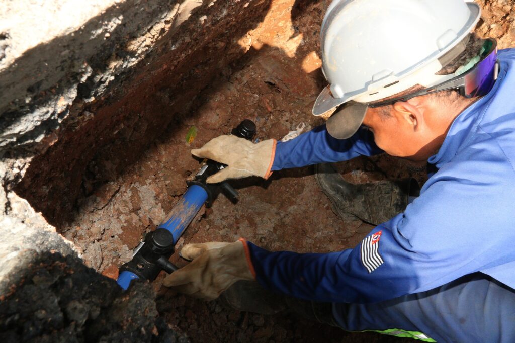 Um trabalhador da construção civil está consertando um cano em uma vala. A imagem mostra um trabalhador usando um capacete de segurança branco, óculos de proteção espelhados e um macacão azul, com luvas de couro bege nas mãos. Ele está ajoelhado em uma vala, com terra marrom e pedras ao redor. Ele está segurando uma ferramenta azul e preta, aparentemente um tipo de chave de cano ou alicate, e trabalhando em um cano azul que está parcialmente enterrado na terra. As luvas e a forma como ele segura a ferramenta indicam que ele está realizando um trabalho de manutenção ou reparo em instalações subterrâneas. O trabalhador tem um físico robusto e a pele escura. Seus óculos de proteção refletem cores vibrantes, sugerindo a luz ambiente. No braço esquerdo do macacão, há um emblema com as cores da bandeira do estado de São Paulo. Ao fundo, percebe-se a profundidade da vala e a textura irregular do solo. A atmosfera da imagem é de trabalho árduo e concentração, com um toque de profissionalismo devido ao equipamento de segurança e uniforme. O humor é prático e focado na tarefa em questão.