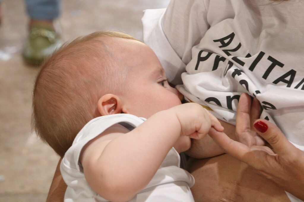 Esta foto captura um momento de carinho e conexão: uma mãe amamentando seu bebê. O bebê, que tem cabelos claros, está aconchegado no colo dela, e sua mãozinha segura suavemente o dedo da mãe, criando um gesto muito delicado. É uma imagem que transmite muita ternura.