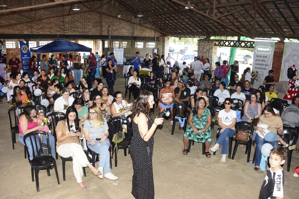 A imagem mostra um evento ou palestra acontecendo em um grande galpão com teto de estrutura metálica e paredes de tijolos aparentes, sugerindo um ambiente rústico. Aqui estão os detalhes principais: Público: Há um grande grupo de pessoas, majoritariamente mulheres, sentadas em cadeiras de plástico pretas organizadas em fileiras, assistindo a uma apresentação. Muitas delas estão com bebês no colo. Apresentação: No centro da imagem, de costas para a câmera, uma mulher com vestido longo estampado de poá (bolinhas) parece estar palestrando ou mediando o encontro, segurando um microfone ou dispositivo. Contexto: O ambiente parece ser um evento familiar ou de apoio à maternidade/saúde, dado que muitas mães estão amamentando ou cuidando de crianças. Há balões, banners de patrocinadores como o "Lions Club" e "Unimed", e até uma pessoa vestida de Homem-Aranha ao fundo. Clima: O tom da reunião é informal e comunitário, com as pessoas atentas, interagindo entre si ou olhando para a frente. Em primeiro plano, no canto inferior direito, uma criança caminha de costas, vestindo uma camisa com a marca "Venum".