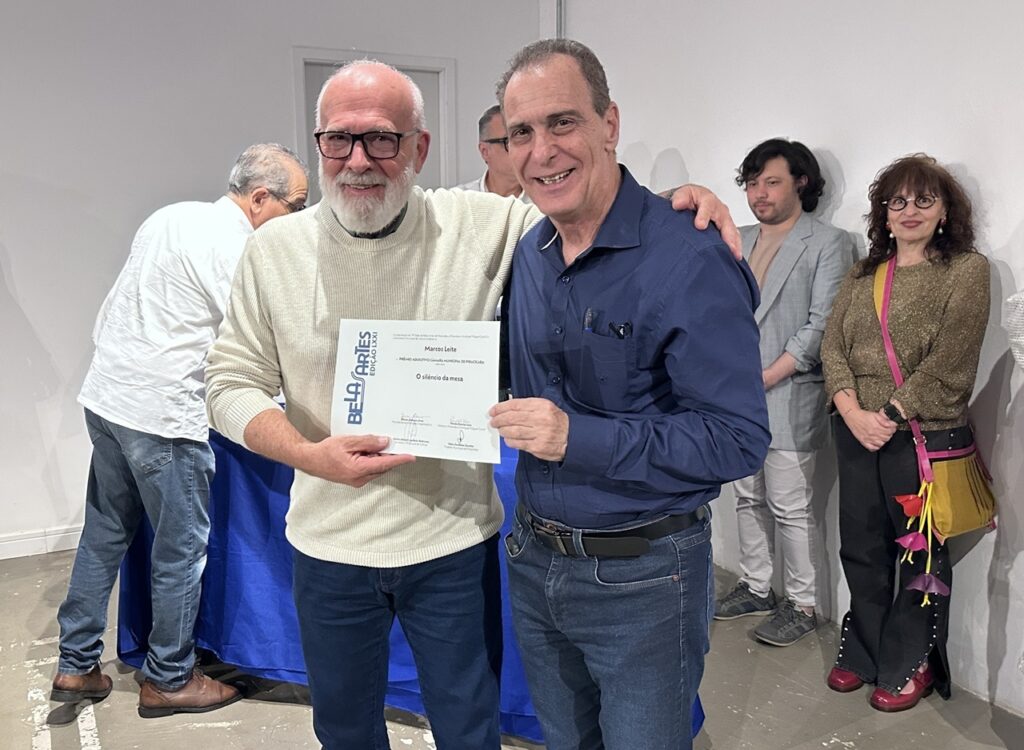 A imagem mostra dois homens posando para uma foto durante uma cerimônia de premiação. Em primeiro plano: O homem à esquerda, de óculos e barba branca, veste um suéter bege e segura um certificado da "Bela Artes". O homem à direita, vestindo uma camisa azul social, está com o braço sobre o ombro do colega, sorrindo para a câmera. O prêmio: O certificado que eles seguram menciona o nome "Marcos Leite" e o título "O silêncio da mesa", além de citar o "Prêmio Aquisitivo Câmara Municipal de Piracicaba". Cenário: O ambiente parece ser um espaço cultural ou galeria, com paredes brancas e uma mesa coberta com uma toalha azul ao fundo. Há outras pessoas na cena: um homem de camisa branca à esquerda e um casal (um homem de paletó cinza e uma mulher com uma bolsa colorida) à direita.