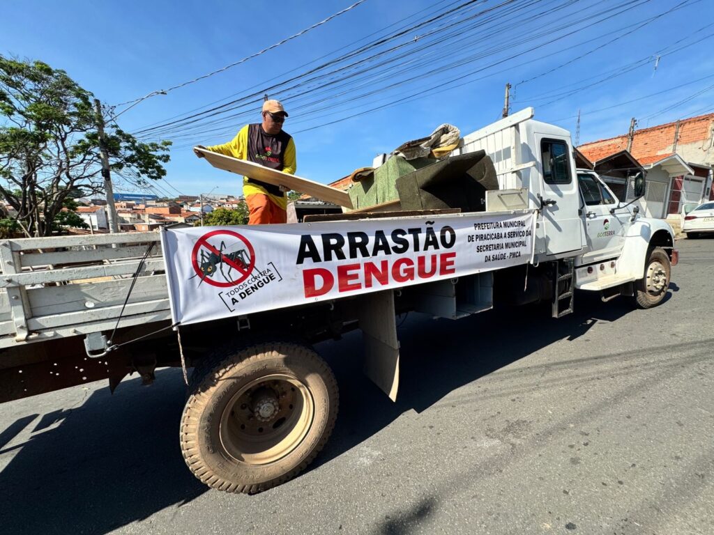 A imagem mostra um caminhão de coleta estacionado em uma rua residencial ensolarada. Na carroceria, há móveis e outros objetos volumosos, enquanto um trabalhador usa luvas, boné, óculos escuros e uniforme amarelo e laranja para carregar uma tábua. Uma faixa no veículo anuncia “ARRASTÃO DENGUE” e traz a mensagem “Todos contra a dengue”. Também informa que a ação é realizada pela Prefeitura Municipal de Piracicaba, por meio da Secretaria Municipal da Saúde. Ao fundo, aparecem casas, árvores, muitos fios elétricos e um céu azul.