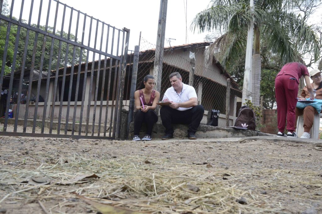 A imagem mostra um grupo de pessoas reunido ao ar livre, em frente a uma casa, atrás de um portão de grades metálicas e uma cerca de arame. Algumas pessoas estão sentadas em cadeiras, enquanto outras permanecem em pé. Elas seguram pranchetas e parecem participar de uma atividade de atendimento, reunião ou ação comunitária. O local tem bastante vegetação, árvores e plantas ornamentais, além de um chão de terra coberto por folhas secas.