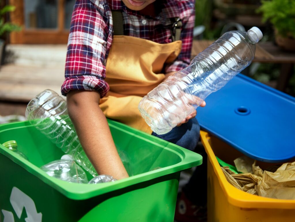 Na imagem, vemos uma pessoa vestindo camisa xadrez e avental enquanto organiza materiais recicláveis em um ambiente externo, como um quintal ou jardim. Ela segura uma garrafa plástica transparente e a coloca em um recipiente verde já cheio de outras garrafas semelhantes. Ao lado, há uma lixeira amarela com tampa azul, destinada a outro tipo de resíduo, possivelmente papel ou plástico. A cena transmite um gesto de responsabilidade ambiental, destacando a importância da separação correta dos materiais para promover a reciclagem, reduzir o desperdício e incentivar práticas sustentáveis no dia a dia.