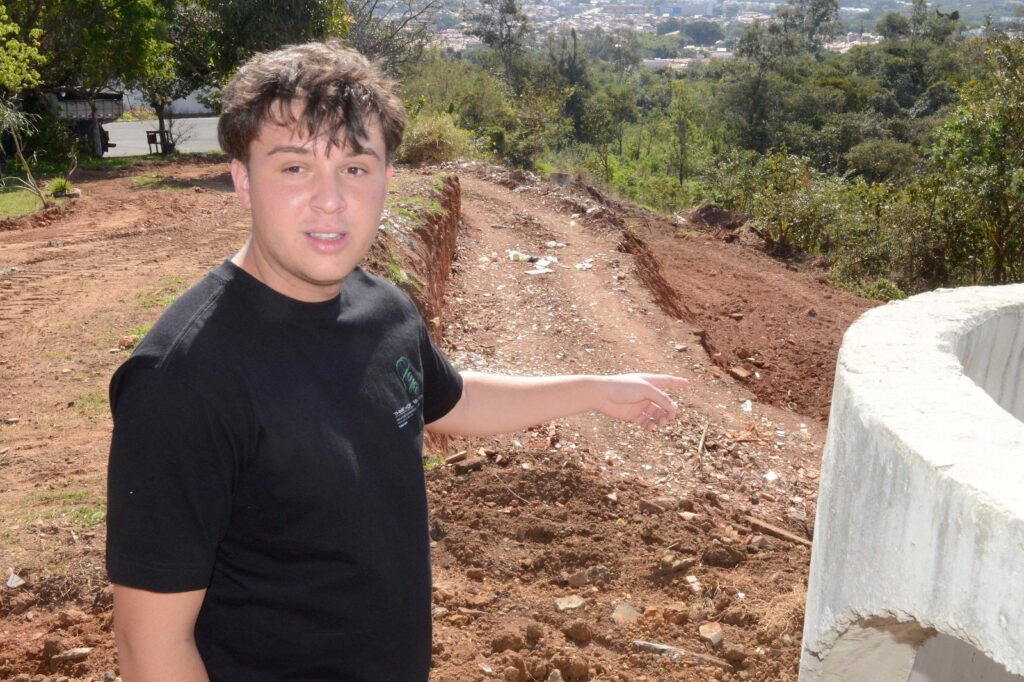 Na foto, observa-se uma pessoa em um terreno em obras ou recentemente escavado, caminhando sobre um caminho de terra solta marcado por rastros de pneus. Ela aponta para a área, indicando algum aspecto da intervenção realizada. Em primeiro plano, há uma estrutura circular de concreto, possivelmente parte de um sistema de drenagem ou infraestrutura em implantação. Ao fundo, aparecem árvores, vegetação e a vista distante de uma cidade, sugerindo que se trata de uma inspeção ou explicação sobre um projeto de desenvolvimento urbano.