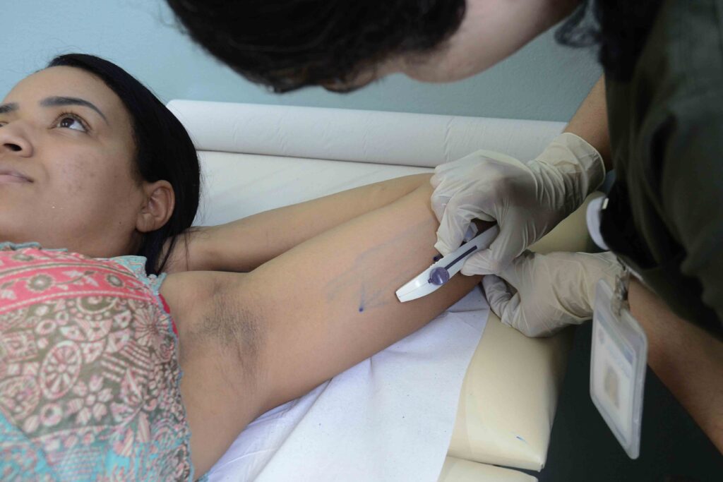 A imagem mostra uma profissional de saúde realizando um exame em uma mulher. A mulher está deitada em uma maca, enquanto a profissional, usando luvas e máscara, para fazer um procedimento.
