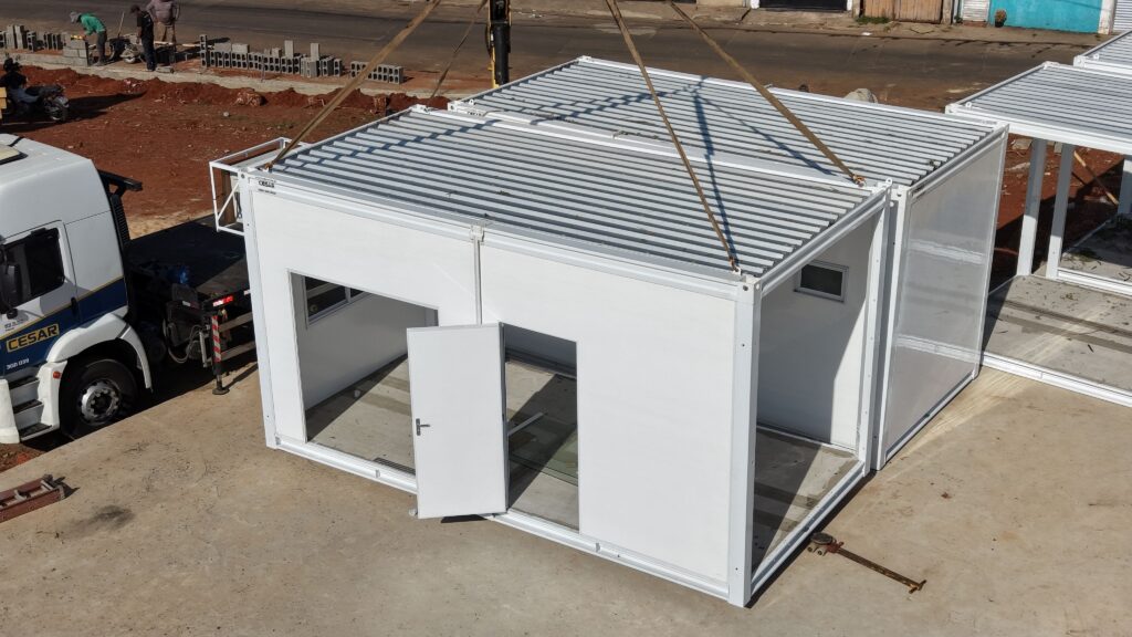 Uma imagem aérea captura o momento preciso em que um guindaste suspende uma estrutura modular pré-fabricada de cor branca, que está sendo posicionada em um canteiro de obras movimentado durante o dia. A composição da cena é focada na estrutura modular central, que domina o quadro, sendo mantida no ar por cabos de aço presos a um guindaste. A perspectiva em ângulo elevado destaca a geometria retangular e industrial da construção, que possui paredes brancas e um telhado metálico corrugado. Ao lado, um caminhão de carga aguarda ou auxilia na operação logística. A cena transmite uma sensação de progresso técnico e eficiência, com uma organização clara entre os elementos de transporte e o objeto modular, demonstrando um processo típico de montagem de canteiros de obras modernos ou habitações modulares. O objeto principal é uma unidade pré-fabricada, composta por painéis metálicos brancos lisos, apresentando uma porta aberta, janelas embutidas e uma estrutura reforçada de metal que sugere durabilidade e facilidade de transporte. A estrutura tem um design limpo e utilitário, com acabamentos industriais, exibindo um telhado que reflete a luz solar. A unidade parece ser composta por dois módulos interconectados, e a sua superfície branca contrasta fortemente com o solo terroso do canteiro de obras, destacando sua natureza como um elemento artificial sendo integrado a um ambiente de construção. A imagem é uma fotografia digital de alta resolução, tecnicamente executada para documentar a precisão da engenharia civil e o trabalho logístico. A nitidez da imagem permite observar detalhes como as dobradiças da porta, a textura do telhado corrugado e o reflexo do ambiente nas superfícies brancas. A escolha do enquadramento, vindo de cima, é fundamental para fornecer uma visão clara da logística do levantamento de peso e da precisão necessária para manusear tais estruturas. O estilo é puramente documental e informativo, capturando o realismo técnico de uma operação de construção com foco na clareza e na escala. O cenário é um canteiro de obras aberto, com o solo de terra batida sob um céu claro e ensolarado. A iluminação é natural e direta, proveniente do topo, criando sombras curtas e bem definidas sob a estrutura, o que reforça sua tridimensionalidade e massa. O ambiente ao redor inclui elementos típicos de construção, como blocos de concreto e veículos, que fornecem contexto à atividade em curso. O clima geral é de produtividade e funcionalidade, refletindo uma atmosfera de construção civil onde cada componente é cuidadosamente posicionado para maximizar a eficiência da montagem no local.