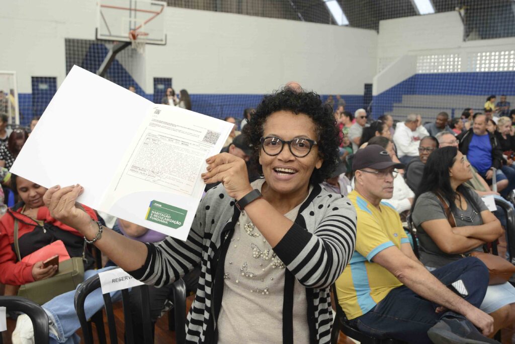 A imagem mostra uma mulher sorridente segurando um documento aberto da Secretaria Municipal de Habitação e Regularização Fundiária, exibindo-o com orgulho em um evento público realizado em um ginásio. Ao fundo, várias pessoas estão sentadas, indicando uma cerimônia de entrega de títulos ou registros de propriedade. O ambiente é descontraído e comunitário, com clima de celebração e conquista pessoal.