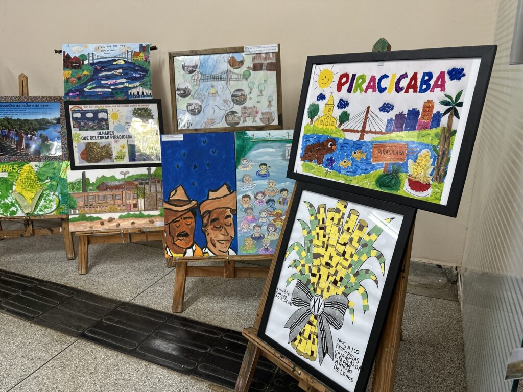 A imagem mostra uma exposição de trabalhos artísticos sobre Piracicaba, organizada em cavaletes e quadros em uma área interna. Em destaque, há uma obra colorida com a palavra “PIRACICABA”, representando o rio, peixes, uma ponte, prédios, uma igreja e elementos da paisagem local. Abaixo, aparece um mosaico de uma espiga de milho, com a inscrição “Mosaico feito pelas crianças da ...”. Ao fundo, há pinturas e colagens com o rio Piracicaba, pontes, construções históricas, paisagens rurais, pessoas nadando e figuras ligadas à cultura da cidade. Os trabalhos usam cores vivas, materiais variados e parecem ter sido produzidos por crianças ou estudantes.