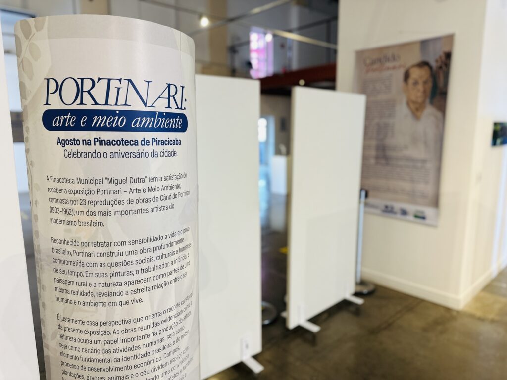 A imagem mostra um cartaz informativo em uma coluna cilíndrica, que anuncia a exposição "Portinari: Arte e Meio Ambiente" na Pinacoteca Municipal "Miguel Dutra", em Piracicaba. Conteúdo do cartaz: O texto explica que a mostra reúne 23 reproduções de obras de Cândido Portinari, um dos maiores nomes do modernismo brasileiro, destacando como o artista retratava com sensibilidade a relação entre o povo brasileiro, o trabalho e o ambiente rural. Cenário: Ao fundo, vê-se o interior da galeria com divisórias brancas e um pôster maior na parede com uma foto de Cândido Portinari, indicando que o espaço está organizado para receber os visitantes da exposição. Parece ser uma homenagem muito bonita ao aniversário da cidade através da arte!
