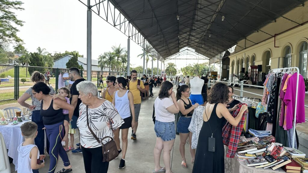 Um grupo diversificado de pessoas passeia por uma feira ao ar livre sob uma estrutura coberta, explorando barracas de roupas e livros.
A cena retrata um dia ensolarado com pessoas de todas as idades circulando em um ambiente que parece ser uma feira ou mercado ao ar livre, possivelmente um evento de artesanato ou usados. A imagem é dominada por uma longa estrutura coberta com um telhado metálico e pilares de sustentação, que se estende por toda a extensão da cena. A luz natural entra pelas laterais abertas, iluminando os visitantes e as mercadorias expostas.
No lado direito da imagem, há uma barraca de roupas com várias peças penduradas em um varal, incluindo vestidos, camisas e blusas em cores vibrantes como rosa choque, estampados e pretos. Abaixo das roupas, há uma mesa com uma pilha de livros. Uma manequim sem cabeça, vestida com shorts azuis, também está exposta. Várias pessoas, predominantemente mulheres, estão próximas à barraca, examinando as roupas e os livros. Uma mulher em um vestido preto longo está olhando para uma camisa xadrez.
No lado esquerdo da imagem, uma mesa menor está coberta com uma toalha branca e expõe alguns itens, como xícaras e recipientes. Há um menino pequeno com um colete cinza olhando para a mesa. Mais adiante, um grupo de pessoas, incluindo adultos e crianças, caminha pelo espaço. Uma mulher carrega uma criança nas costas. Outras pessoas passeiam mais ao fundo, em direção à saída da estrutura coberta. A arquitetura do prédio principal, com arcos e janelas, é visível ao fundo.
O ambiente geral é de movimento e interação social. A atmosfera é casual e descontraída, com pessoas aproveitando o dia para comprar, vender e socializar. O humor parece ser positivo e animado, típico de um evento comunitário. As cores são variadas, com destaque para as roupas coloridas na barraca e os tons verdes da vegetação visível nas áreas externas. As sombras projetadas pela estrutura coberta criam um contraste com as áreas iluminadas pelo sol, adicionando profundidade à imagem.