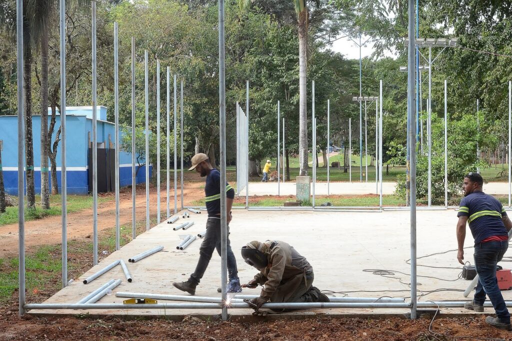 Nesta imagem, vemos três homens trabalhando na construção ou montagem de uma estrutura metálica em uma área aberta, possivelmente um parque. O trabalho: O trabalhador central está ajoelhado, usando uma máscara de solda e avental de proteção, concentrado em soldar uma das hastes verticais da estrutura. Há faíscas visíveis no ponto de solda. Os outros trabalhadores: À esquerda, um homem de boné caminha pela estrutura, carregando algum equipamento ou material. À direita, outro homem, também usando um colete refletivo, observa o trabalho ou se prepara para ajudar. O local: A estrutura é formada por vários postes metálicos finos fixados em uma base de concreto nivelada. Ao fundo, há muitas árvores, uma área gramada e uma construção azul com detalhes em branco. O ambiente parece ser uma obra ao ar livre durante o dia.