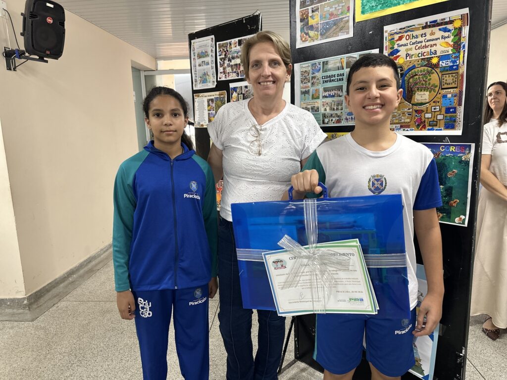 A foto mostra duas crianças e duas mulheres em um ambiente escolar, diante de painéis com trabalhos sobre Piracicaba. As crianças usam uniformes esportivos azuis da Prefeitura de Piracicaba. O menino segura uma grande pasta ou caixa azul transparente, decorada com uma fita prateada e um certificado de reconhecimento. Ao fundo, aparecem cartazes com imagens, desenhos e textos sobre a cidade, incluindo referências ao rio Piracicaba e ao projeto “Olhares que celebram Piracicaba”. Todos posam sorrindo para a foto.