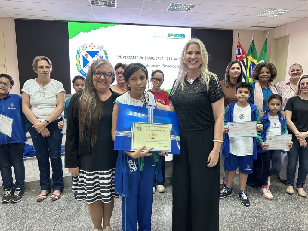 A foto mostra um grupo de crianças e adultos reunidos em uma cerimônia de reconhecimento em um ambiente institucional. Ao centro, uma menina segura uma pasta azul grande com um certificado; outras crianças também exibem certificados. Ao fundo, há uma tela com o brasão e a identidade visual de Piracicaba, além do texto “Aniversário de Piracicaba – 259 anos” e uma referência a “Olhares que celebram Piracicaba”. À direita, aparecem as bandeiras do estado de São Paulo, do Brasil e de Piracicaba. Todos posam diante da câmera, e algumas crianças vestem uniformes azuis da Prefeitura de Piracicaba.