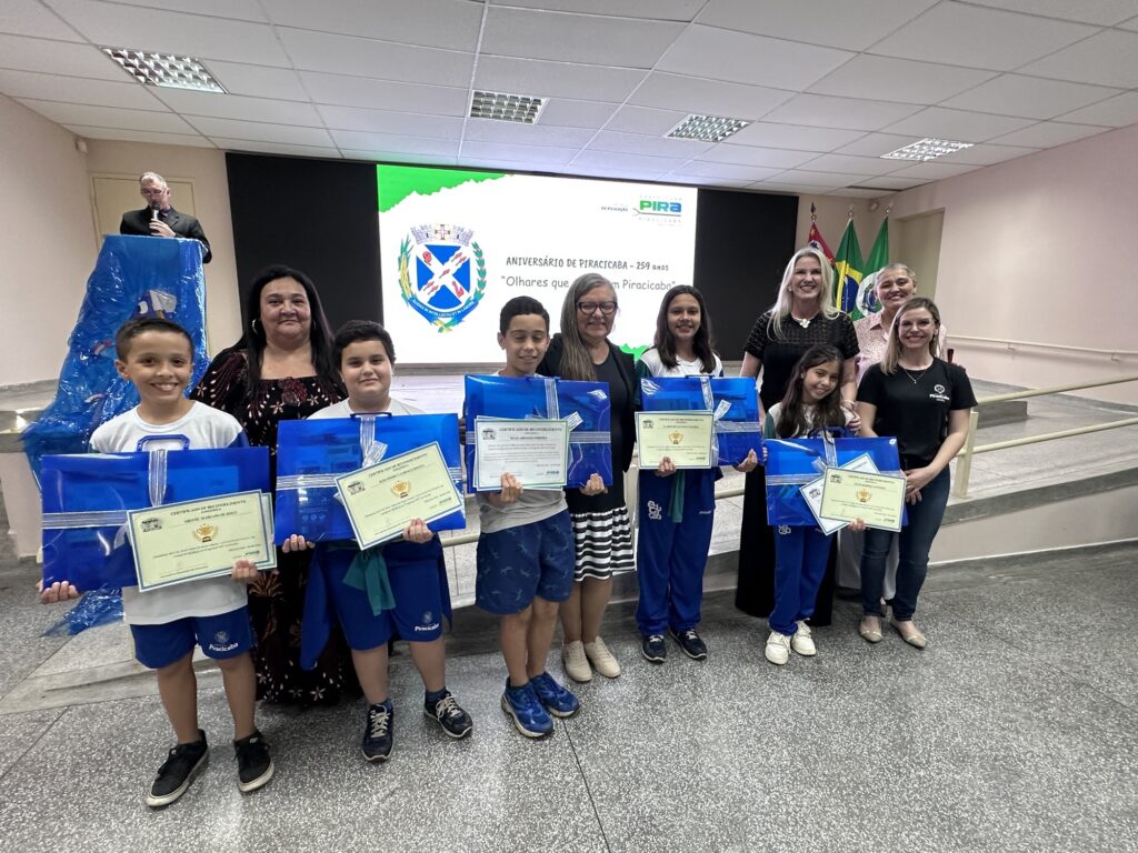 A foto mostra um grupo de crianças e adultos em uma cerimônia de reconhecimento, realizada em um auditório. As crianças seguram pastas azuis decoradas com fitas prateadas e certificados. Ao fundo, um telão exibe o brasão de Piracicaba e os dizeres “Aniversário de Piracicaba – 259 anos” e “Olhares que celebram Piracicaba”. Também aparecem as bandeiras do estado de São Paulo, do Brasil e de Piracicaba. À esquerda, um homem fala ao microfone atrás de um púlpito coberto de azul.