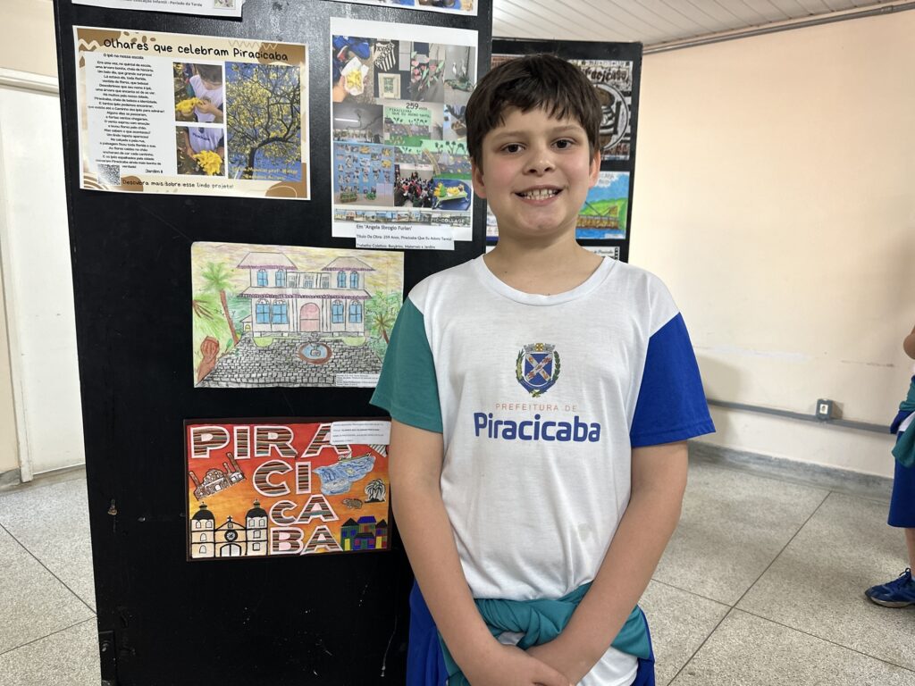 A foto mostra um menino sorrindo, em primeiro plano, usando uma camiseta da Prefeitura de Piracicaba. Ele está diante de um painel escolar preto com trabalhos artísticos sobre a cidade. Entre as obras, aparecem um cartaz intitulado “Olhares que celebram Piracicaba”, uma colagem sobre os 259 anos de Piracicaba, desenhos de prédios históricos, paisagens, o rio e a palavra “PIRACICABA” em destaque. O ambiente parece ser uma escola ou espaço de exposição, com outras crianças parcialmente visíveis ao fundo.