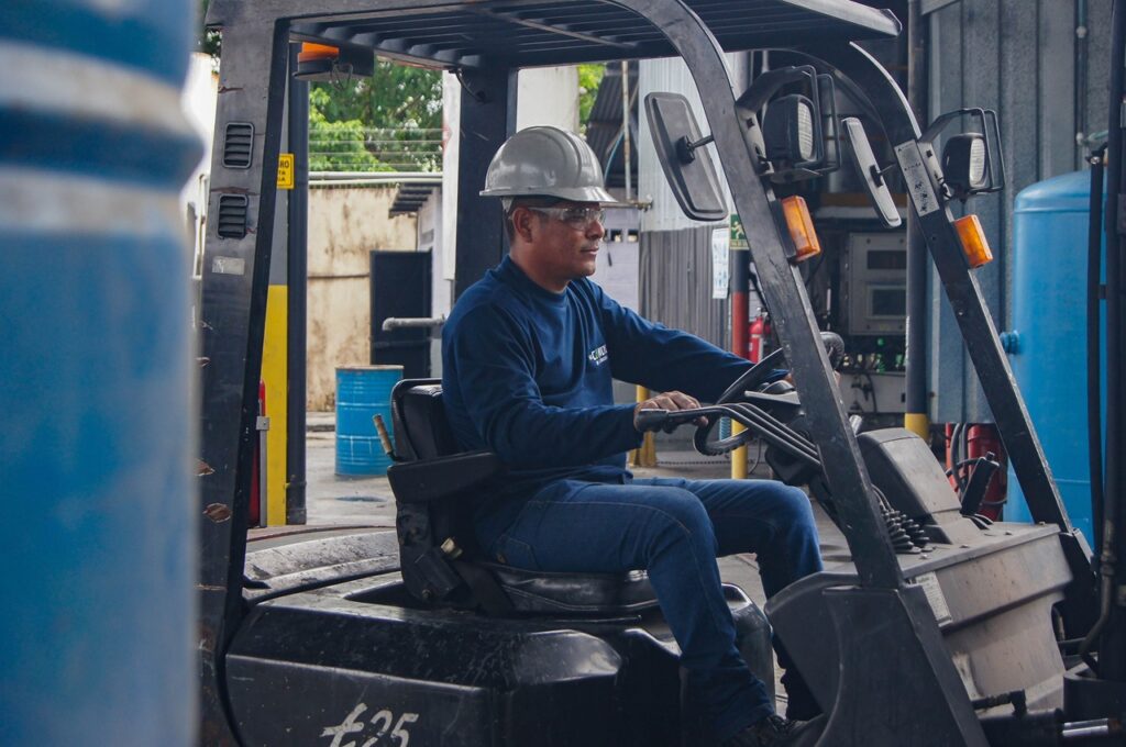 A imagem mostra um trabalhador operando uma empilhadeira em um ambiente industrial. Ele usa capacete de segurança, óculos de proteção e uniforme azul, demonstrando boas práticas de segurança no trabalho. O cenário inclui tambores azuis e estruturas metálicas, típicos de um armazém ou área de carga e descarga.