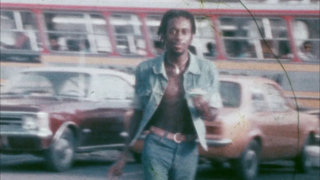 A imagem tem uma estética vintage, lembrando uma fotografia antiga ou um frame de filme de décadas passadas. O protagonista: Um homem negro com dreadlocks, óculos e um colar, caminha em direção à câmera. Ele veste um colete jeans aberto sobre o peito nu e uma calça jeans com um cinto de fivela marcante. Ele tem um olhar calmo e direto para a lente. O cenário: Ele está em uma rua movimentada. Ao fundo, percebem-se veículos clássicos, como um carro escuro à esquerda e um modelo alaranjado à direita, típicos de modelos brasileiros dos anos 70 ou 80 (como Opala ou Chevette). O estilo: A imagem possui marcas de desgaste e textura granulada, como se fosse uma película de cinema envelhecida, o que dá uma atmosfera nostálgica e autêntica à cena urbana capturada.