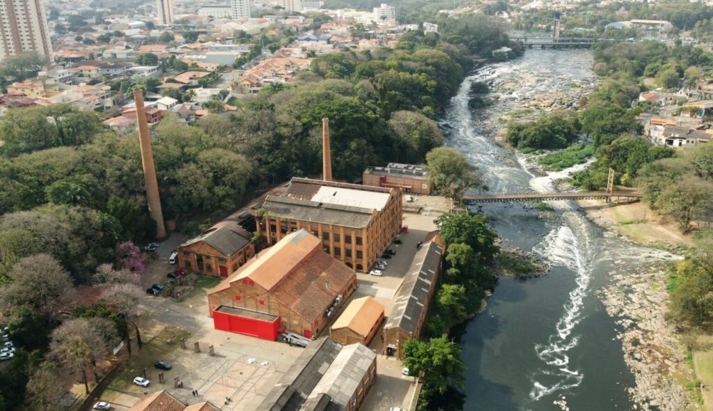 Vista aérea de uma antiga área industrial junto a um rio, integrada a uma paisagem urbana e de mata ribeirinha. Composição geral: prédio industrial de tijolos no centro da imagem, com vários blocos conectados formando um complexo; à direita corre um rio com trechos de corredeiras e margens rochosas; ao fundo, bairros residenciais e alguns prédios altos. Edifícios e materiais: telhados inclinados de cerâmica e chapas metálicas envelhecidas; fachadas de tijolo aparente com janelas regularmente espaçadas; um dos galpões tem uma grande porta ou painel pintado de vermelho vivo, que se destaca no conjunto. Chaminés: duas chaminés altas e finas de alvenaria (uma bem visível à esquerda do complexo) evocam uso industrial passado — servem como elemento vertical marcante na cena. Pátios e circulação: áreas abertas pavimentadas e pátios entre os prédios, algumas vagas de estacionamento com carros, e caminhos de serviço que conectam os blocos; presença de pequenas áreas gramadas e árvores esparsas próximas aos edifícios. Vegetação e entorno natural: faixa densa de árvores entre o complexo e o bairro, formando um corredor verde ribeirinho; margem do rio com arbustos e pedras expostas; no lado oposto do rio, vegetação mais contínua e poucas construções. Rio e infraestruturas: água escura com ondulações e espumas em trechos rasos; uma ponte metálica estreita cruza o rio próxima ao complexo, ligando as duas margens; ao longe há outra ponte ou barragem. Cores e luz: tons terrosos predominam (tijolo, telha), contraste com o vermelho da porta e o verde da vegetação; luz suave, provavelmente final de manhã ou tarde com sombras discretas. Atmosfera e impressão: sensação de histórico industrial bem preservado ou adaptado, integrado à cidade e ao ambiente natural — cenário que combina memória industrial, uso atual e valorização do espaço ribeirinho.
