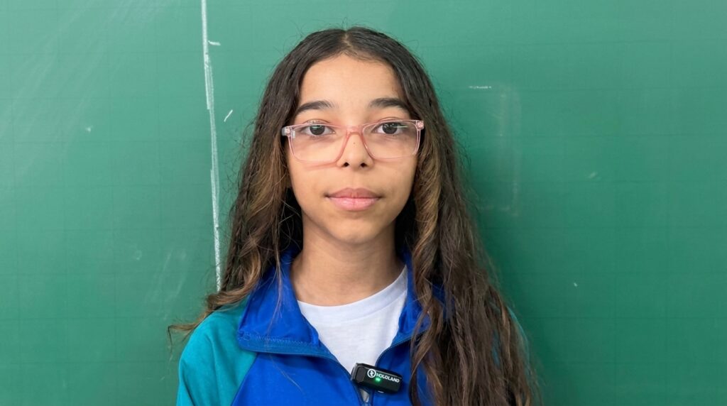 Nesta imagem, aparece uma menina em frente a um quadro verde, vestida com uma jaqueta azul e turquesa sobre uma camiseta branca. Ela usa óculos cor-de-rosa e tem cabelos longos e castanhos. O quadro atrás está limpo, com leves marcas de grade e um traço branco de giz à esquerda, reforçando o contexto de sala de aula.