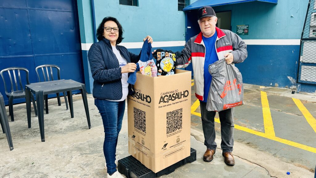 A imagem mostra duas pessoas participando de uma Campanha do Agasalho, uma ação solidária para arrecadar roupas e cobertores. Elas estão ao lado de uma grande caixa de papelão identificada com o slogan “Doe com amor. Faz bem fazer o bem.”, colocando peças de roupa dentro dela. O ambiente é externo, com paredes azuis e algumas cadeiras plásticas ao fundo, sugerindo um ponto de coleta comunitário.