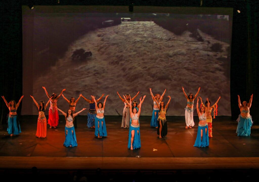Na imagem, um grupo de dezesseis dançarinas se apresenta em um palco, todas vestindo trajes coloridos com saias esvoaçantes e tops decorativos, típicos da dança do ventre. Elas estão dispostas em formação, com os braços erguidos em movimentos sincronizados, transmitindo energia e harmonia. Ao fundo, um grande telão projeta a imagem de um rio turbulento, criando um contraste dramático entre a delicadeza da dança e a força da natureza.