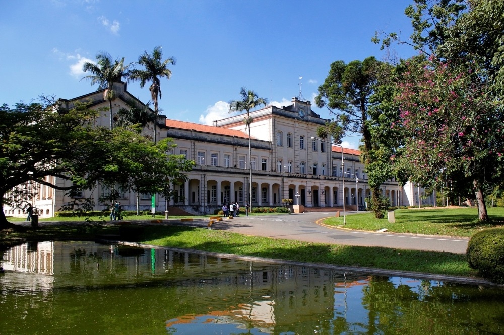 Grande edifício histórico de fachadas claras visto em um dia ensolarado, com jardim e lago em primeiro plano. Arquitetura: prédio amplo em estilo clássico/colonial, com dois pavimentos e corpo central levemente saliente; varandas com arcos no térreo e janelas retangulares no andar superior. Às vezes há ornamentos no topo do corpo central (possivelmente um pequeno frontão com detalhe circular). Telhado de telhas vermelhas. Fachada e cores: tons creme/claro com molduras e pilares discretos; aparência bem conservada e institucional. Vegetação: palmeiras altas perto da fachada, árvores grandes à direita (uma com flores rosadas) e um grande árvore de copa larga à esquerda que projeta sombra sobre o gramado. Lago/reflexo: espelho d’água calmo em primeiro plano que reflete parte do prédio, do céu e da vegetação, acrescentando profundidade à cena. Caminhos e área externa: rua ou circulação em curva à frente do edifício, bancos e pequenos canteiros bem cuidados; gramados amplos e arborizados. Pessoas: pequeno grupo de pessoas próximas à entrada central — atuando como escala humana, sem detalhes identificáveis. Céu e iluminação: céu azul com poucas nuvens; iluminação forte, cores vivas e sombras definidas, sugerindo final de manhã ou início de tarde.