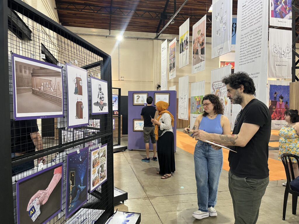 A imagem mostra uma exposição de ilustrações e histórias em quadrinhos montada em painéis suspensos e grades metálicas, dentro de um salão com teto de madeira. À esquerda, há obras numeradas, incluindo desenhos de um prédio identificado como “PIRILÕES”, personagens em uma porta e um robô. À direita, os painéis apresentam ilustrações coloridas, caricaturas e textos em inglês, com nomes de artistas como Karma Leão, André Dahmer e Thiago Limo. Algumas obras abordam tecnologia, robôs, o futuro e situações do cotidiano com humor. Visitantes circulam pelo espaço observando os trabalhos; em primeiro plano, uma mulher de blusa azul segura uma prancheta e parece fazer anotações.