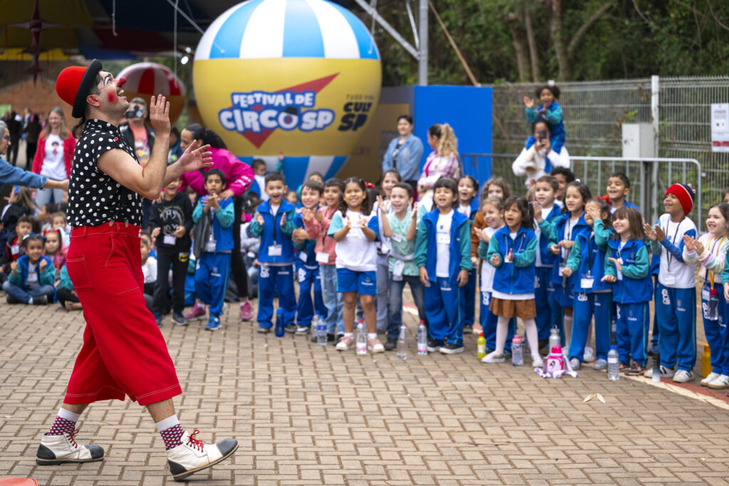 A imagem mostra um palhaço animando um grupo de crianças em um evento ao ar livre, identificado como o Festival de Circo SP. O artista usa roupas coloridas — calça vermelha, camisa preta com bolinhas brancas, chapéu vermelho e nariz de palhaço — enquanto interage com o público infantil, que parece encantado e sorridente. As crianças, vestidas com uniformes escolares azuis e brancos, assistem e aplaudem entusiasmadas. Ao fundo, há um grande balão decorativo com o logotipo do festival e pessoas acompanhando a apresentação.