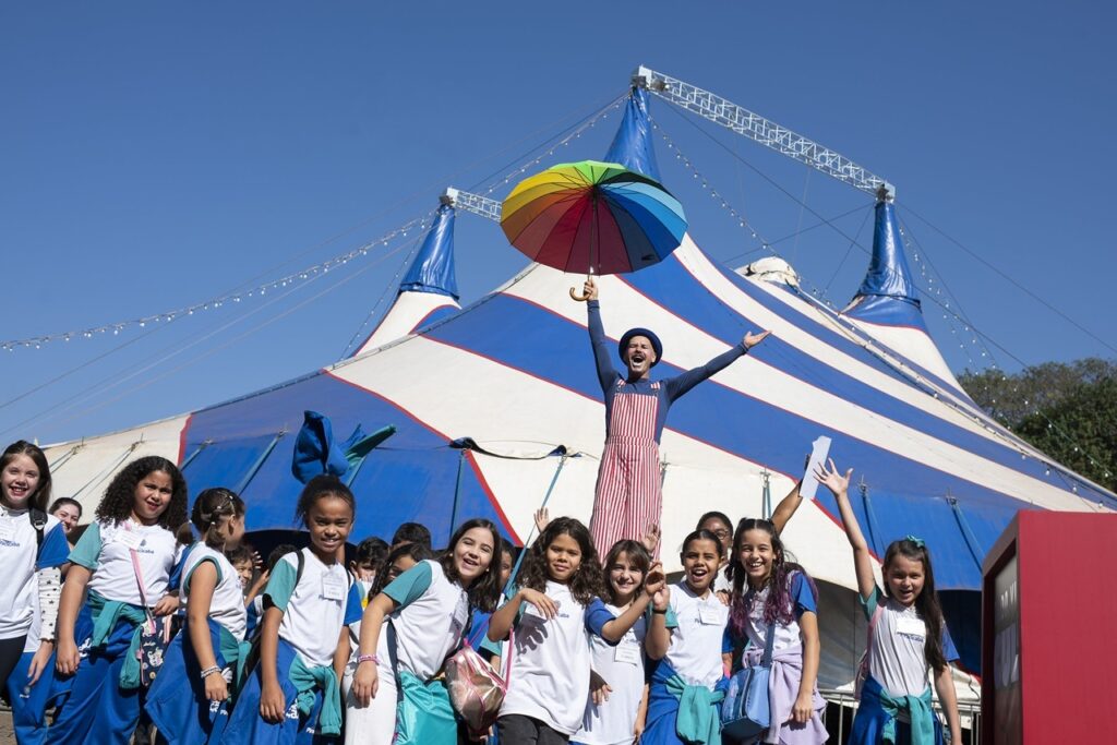 A imagem mostra um grupo de crianças sorrindo e posando em frente a um grande circo com lona listrada em azul, branco e vermelho. Elas usam camisetas brancas com detalhes azuis e carregam mochilas ou bolsas. No centro, um palhaço está sobre uma plataforma, usando chapéu azul e macacão listrado vermelho e branco. Ele segura um guarda-chuva colorido e levanta os braços, enquanto as crianças comemoram ao redor. O céu está azul e ensolarado, criando um clima alegre e festivo.