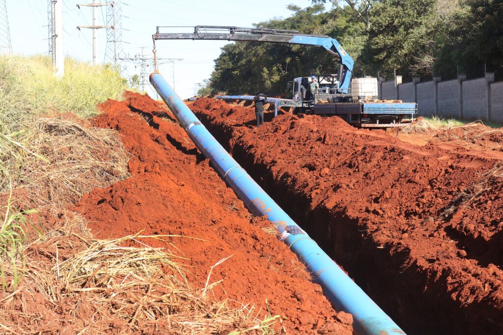 A cena retrata uma obra de infraestrutura em andamento, focada na instalação de um grande tubo azul em uma vala cavada em solo de terra vermelha. A perspectiva está ligeiramente inclinada, criando uma sensação de profundidade e movimento. O elemento principal é o tubo azul, de diâmetro considerável, que atravessa a vala na diagonal, avançando da parte inferior direita para a superior esquerda da imagem. Ele repousa sobre o fundo da vala e em algumas partes é sustentado por montanhas de terra. O tubo apresenta algumas marcas e um adesivo com código de barras ou identificação na parte central direita. A vala é profunda e as paredes de terra vermelha são visíveis, com alguns tufos de grama seca e palha espalhados nas bordas superiores e ao redor. A cor vibrante da terra contrasta fortemente com o azul do tubo e com o verde da vegetação ao fundo. No fundo, um caminhão munck azul e branco está posicionado. O braço hidráulico do munck está erguido e conectado ao tubo azul, indicando que o mesmo está sendo posicionado ou manobrado. Há duas pessoas na área do caminhão: uma vestindo um capacete azul e uma camisa clara, observando a operação, e outra, mais ao fundo, de preto, com a cabeça levantada, possivelmente direcionando o trabalho. A presença de um tanque branco de grande porte (possivelmente de água ou outro material) no caminhão é notável. O cenário ao redor inclui uma fileira de postes de alta tensão metálicos e estruturas de energia elétrica à esquerda, com vegetação rasteira e arbustos. À direita, uma mureta de blocos de concreto cinzas se estende, separando a área da obra de uma área com mais vegetação e árvores ao fundo, sob um céu azul claro e sem nuvens. A composição utiliza a diagonal do tubo e da vala para guiar o olhar do espectador pela cena, criando um dinamismo interessante. A luz do sol parece forte, pois as sombras não são muito pronunciadas, indicando um dia claro. Os detalhes como a textura da terra, a grama seca e as marcações no tubo adicionam realismo à imagem. As cores predominantes são o vermelho intenso da terra, o azul vibrante do tubo e do caminhão, e os tons de verde da vegetação.