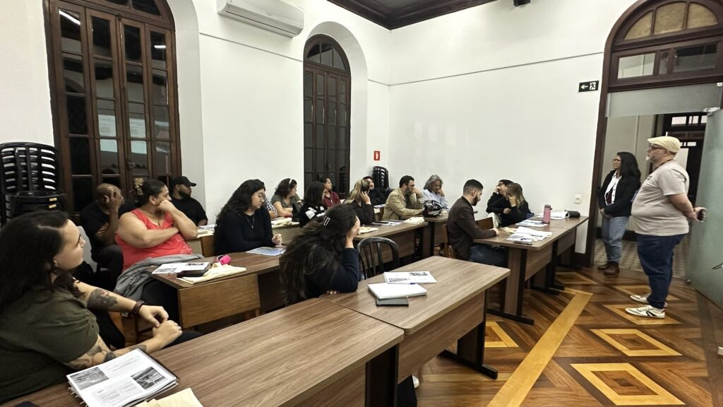 Sala de aula/treinamento com cerca de 16 pessoas participando de uma atividade presencial: Disposição: fileiras de carteiras e mesas de madeira ocupadas por participantes que olham para a frente e conversam entre si; alguns têm cadernos, apostilas e canetas à frente. Participantes: mistura de homens e mulheres adultos de idades variadas; expressão atenta ou conversadora, postura relaxada. Facilitadores: duas pessoas em pé na frente à direita — um homem de camiseta clara, boné e óculos, com as mãos atrás; e uma mulher de blazer escuro. Eles parecem conduzir ou observar a atividade. Ambiente arquitetônico: sala com paredes brancas altas, portas/janelas de madeira com vitrôs e ar-condicionado instalado acima; piso de madeira com desenho geométrico bem visível. Materiais visíveis: pilha de cadeiras plásticas empilhadas ao fundo à esquerda; papéis, apostilas e garrafas de água sobre as mesas; alguns participantes com mochilas e capacete sobre a mesa. Iluminação e clima: luz artificial clara, ambiente interno organizado e formal — sensação de reunião, oficina ou aula prática. Dinâmica: participantes parecem divididos entre ouvir a frente da sala e discutir entre si; atmosfera séria-moderada, focada em aprendizado ou planejamento.
