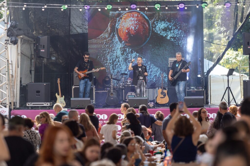 Palco ao ar livre com banda tocando e público em frente, durante um evento diurno. Cena geral: palco centralizado com um telão grande ao fundo mostrando uma imagem (uma bola/objeto circular visto de cima). À frente, três músicos visíveis (guitarrista à esquerda, vocalista ao centro, baixista à direita) e bateria ao fundo. Músicos e instrumentos: guitarras elétricas e acústicas apoiadas no palco; o guitarrista veste camisa preta e óculos escuros, concentra-se no instrumento; o vocalista segura o microfone com a mão direita erguida, gesticulando; o baixista toca com postura firme. Ao fundo, bateria e amplificadores. Iluminação e estrutura: fileira de luzes decorativas e spots coloridos no topo do palco; laterais com caixas de som e estrutura metálica visível; leve fumaça de cena/efeito de luz à direita. Público: muitas pessoas em primeiro plano, sentadas em mesas e em pé, conversando, olhando para o palco e registrando com celulares; ambiente descontraído e animado, típico de festival ou encontro público. Ambiente e clima: evento ao ar livre em dia claro (árvores ao fundo sugerem parque); sensação de confraternização, música ao vivo e atmosfera festiva. Detalhes menores: mesa/laptop à direita do palco, cabos e equipamentos visíveis, banner rosa na frente do palco parcialmente legível, várias garrafas e copos nas mesas do público.