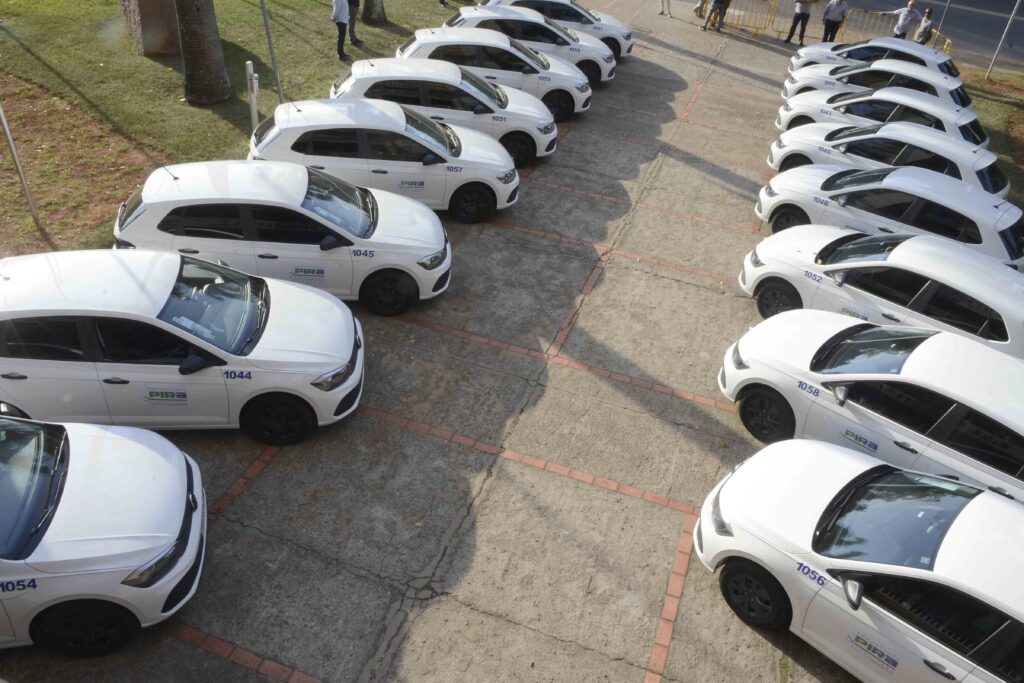 A imagem, vista de cima, mostra muitos carros brancos estacionados em fileiras, aparentemente veículos de serviço identificados com números como 1043, 1044, 1045, 1051, 1053 e 1057. Alguns exibem o logotipo “PIR…” nas portas. Os carros estão em uma área pavimentada com calçamento de pedras e bordas de tijolos vermelhos. À esquerda há uma faixa de gramado com árvores e postes, enquanto, ao fundo, aparecem pessoas próximas a grades metálicas amarelas.