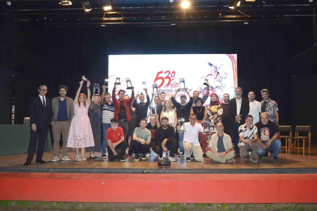 A imagem mostra uma grande foto de grupo em um palco, provavelmente durante um evento ou apresentação. Dezenas de pessoas estão reunidas, algumas em pé e outras sentadas, sorrindo para a câmera; várias levantam os braços em comemoração. Ao fundo, há uma tela iluminada com ilustrações coloridas e letras grandes em vermelho, além de refletores e cabos no teto. O palco tem piso de madeira, cadeiras e uma faixa vermelha na parte da frente.