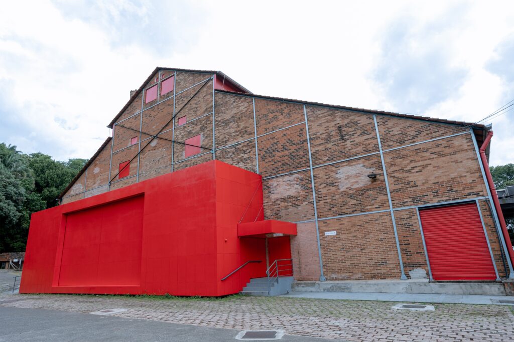 A imagem mostra a fachada de um grande edifício de tijolos aparentes, com arquitetura industrial e telhado inclinado. A parte inferior é marcada por uma estrutura moderna pintada de vermelho, que inclui uma entrada com escadas, uma porta de acesso técnico e uma grande porta metálica de enrolar. As janelas e algumas aberturas estão cobertas por painéis vermelhos, enquanto vigas e reforços metálicos cinzentos atravessam as paredes. Há placas indicando “Entrada técnica” e “Bilheteria”. O prédio fica junto a uma calçada de paralelepípedos, cercado por árvores, sob um céu claro com nuvens.