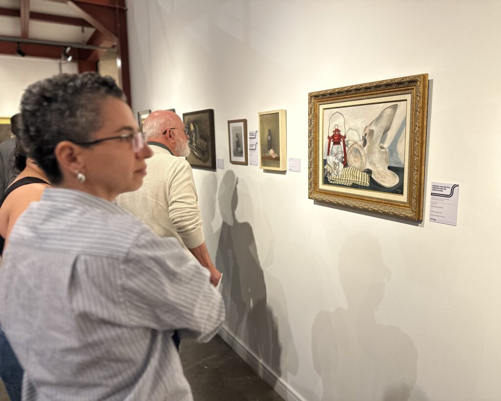 A imagem mostra uma visita a uma exposição de arte em uma galeria ou museu. Em primeiro plano: Vemos de perfil uma pessoa de cabelos curtos, grisalhos e cacheados, usando óculos, que observa atentamente um quadro. Ao fundo: Um homem de barba branca e suéter bege também observa a exposição. Eles estão diante de uma parede branca onde várias obras estão penduradas. A obra em destaque: À direita, há um quadro centralizado, com uma moldura dourada trabalhada. A pintura retrata um objeto que parece ser um osso grande ou uma estrutura óssea, ao lado de uma lanterna vermelha e um pano listrado. Informações adicionais: Ao lado do quadro, há uma ficha técnica detalhando a obra: "Prêmio Aquisitivo Prefeitura de Piracicaba", o título "Ossos do ofício", técnica "Acrílico sobre tela" e dimensões. O ambiente é iluminado e clean, típico de espaços de exibição cultural.