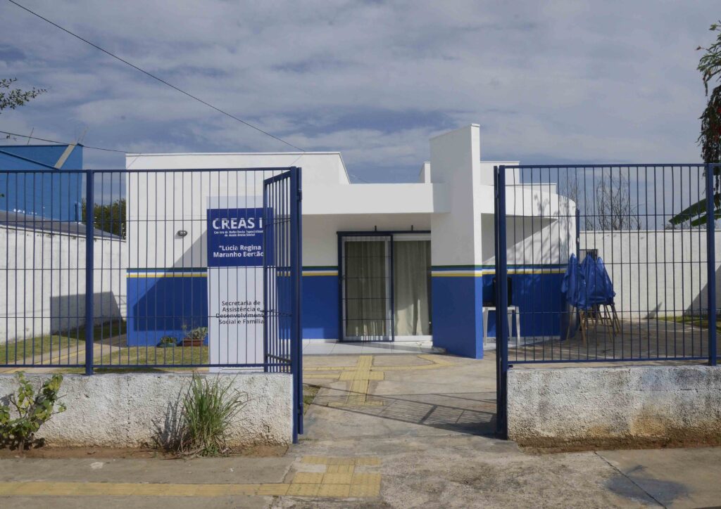 A imagem mostra a fachada de um prédio institucional recém-reformado, identificado como CREAS I – Centro de Referência Especializado de Assistência Social “Lúcia Regina Maranho Bertac”. O edifício é pintado em branco e azul, com uma faixa amarela horizontal, e cercado por um portão metálico azul. O acesso principal tem piso tátil amarelo, indicando preocupação com acessibilidade. O ambiente está limpo e bem cuidado, com plantas próximas ao muro e cadeiras cobertas ao lado direito, sugerindo preparação para um evento ou inauguração.