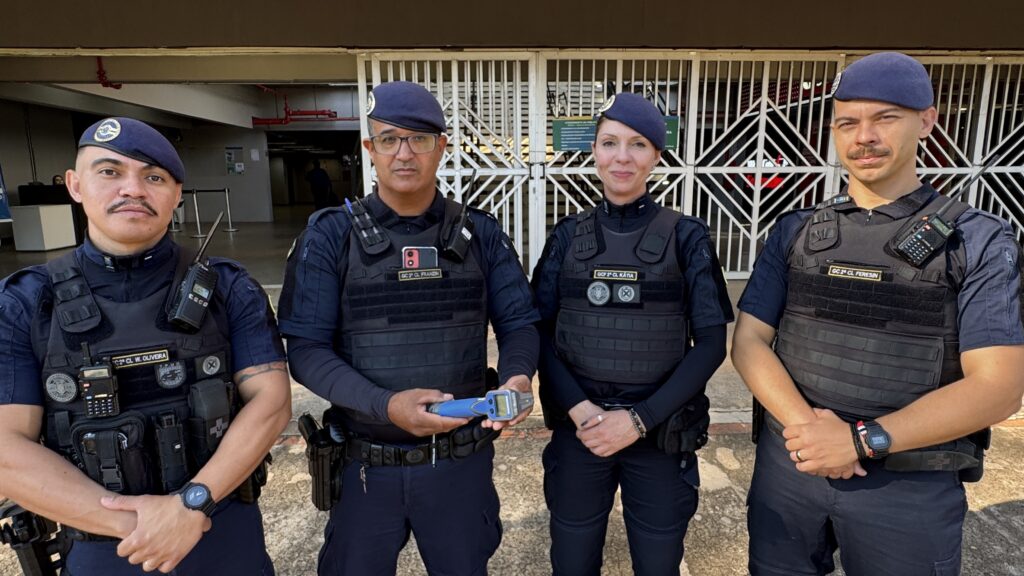 A foto mostra quatro agentes uniformizados, usando boinas azul-marinho, coletes táticos e equipamentos de comunicação. Eles estão em frente à entrada gradeada de um prédio, posando para a câmera. O agente ao centro segura um equipamento azul, enquanto os demais mantêm os braços cruzados ou apoiados na cintura. Há crachás e identificações nos coletes, mas alguns textos estão parcialmente difíceis de ler.