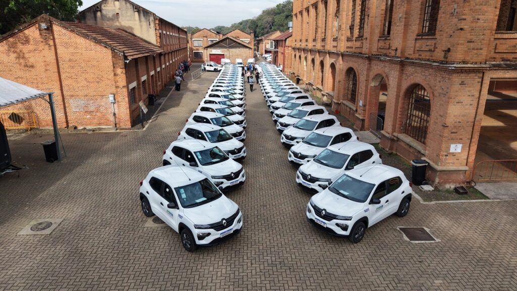 Esta imagem mostra uma grande frota de carros brancos, modelo Renault Kwid, estacionados de forma perfeitamente alinhada em duas colunas paralelas, em um pátio pavimentado com blocos de concreto. Localização: O pátio é ladeado por construções de tijolos aparentes com arcos e janelas grandes, sugerindo que se trata de um complexo histórico ou industrial antigo, possivelmente na cidade de Piracicaba (dado que se consegue ler "Pira" e números de frota nos veículos, como "1107" e "1451"). Organização: A simetria dos carros, todos virados na mesma direção, cria um efeito visual de ordem e abundância. Há um espaço livre no centro entre as duas colunas de veículos. Pessoas: Ao fundo, é possível ver algumas figuras humanas caminhando pelo pátio e observando a frota, dando uma dimensão da escala do local. Atmosfera: O dia parece estar claro e ensolarado. A cena transmite uma sensação de entrega oficial, inspeção de frota ou um evento de renovação de veículos de uma instituição pública ou empresa.
