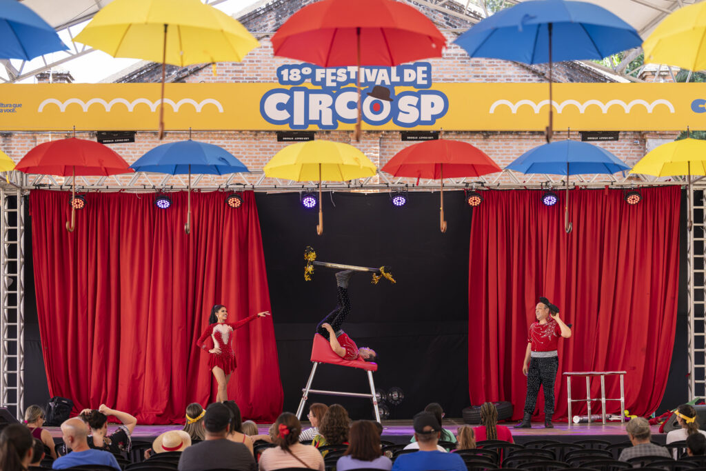 A imagem mostra uma apresentação circense durante o 18º Festival de Circo SP. No palco, decorado com cortinas vermelhas e guarda-chuvas coloridos suspensos, três artistas realizam um número acrobático. Um deles está de cabeça para baixo sobre um aparelho, equilibrando uma barra com os pés; outro, à esquerda, usa um figurino vermelho com detalhes brancos e faz uma pose elegante; e o terceiro, à direita, observa a performance com expressão divertida. O público assiste atentamente, criando um clima alegre e vibrante típico de festivais de circo.