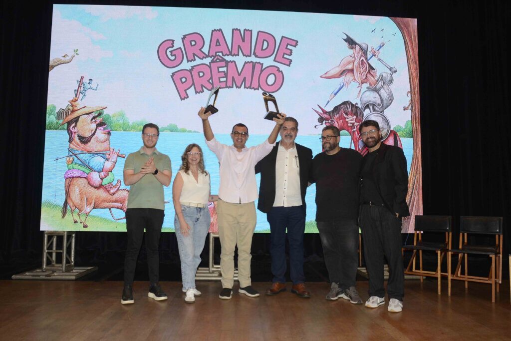 A imagem mostra um grupo de pessoas reunido no palco de um evento, em frente a um grande painel colorido com a palavra “GRAFITARTE” em letras cor-de-rosa. O fundo também apresenta ilustrações de personagens e paisagens. Há homens e mulheres de diferentes idades, alguns usando óculos. Duas pessoas seguram livros ou cadernos abertos acima da cabeça, enquanto outras sorriem e fazem poses para a foto. Todos estão diante de várias cadeiras vazias, sobre um piso de madeira, em um ambiente que parece ser um teatro ou auditório.