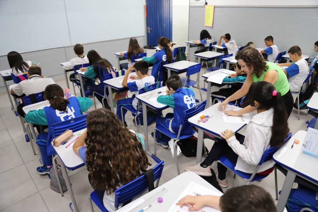 Esta imagem mostra uma sala de aula de ensino fundamental ou médio durante uma atividade. Aqui estão os pontos principais: Ambiente: É uma sala de aula organizada, com mesas individuais dispostas em fileiras. Há uma lousa branca grande à esquerda. Alunos: Os estudantes estão sentados em suas mesas, a maioria vestindo uniformes escolares que parecem ter detalhes em azul, verde e branco (com a palavra "Educa" estampada nas costas de alguns coletes). Eles estão concentrados em tarefas ou exercícios em papéis sobre as mesas. Professora: À direita, uma professora vestindo uma blusa verde clara está em pé, inclinada sobre a mesa de uma aluna, auxiliando-a com o material de estudo. Atmosfera: O clima parece de uma aula comum, com todos os alunos trabalhando silenciosamente em suas atividades. Há materiais escolares como estojos e canetas espalhados pelas mesas.