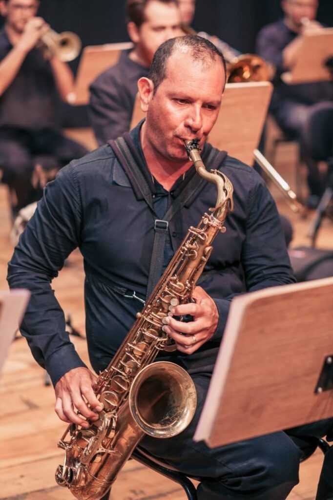 A imagem mostra um músico tocando saxofone em uma apresentação de orquestra. Ele está sentado, usando uma camisa escura e uma correia para apoiar o instrumento dourado. Ao fundo, outros músicos tocam instrumentos de sopro, enquanto estantes com partituras aparecem espalhadas pelo palco.