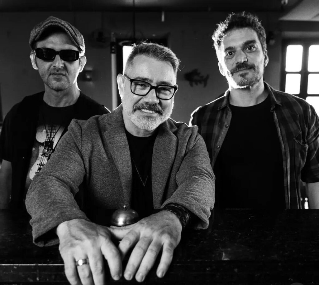 A imagem em preto e branco mostra três homens em um ambiente que parece ser um bar ou restaurante. O homem no centro está mais próximo da câmera, usa óculos, barba grisalha e um blazer sobre camiseta escura. Ele apoia as mãos sobre o balcão, ao lado de uma pequena campainha. À esquerda, há um homem usando boné, óculos escuros e camiseta preta. À direita, aparece um homem de barba curta, vestindo uma camisa xadrez aberta sobre uma camiseta. Ao fundo, há janelas e iluminação forte, criando um contraste marcante.