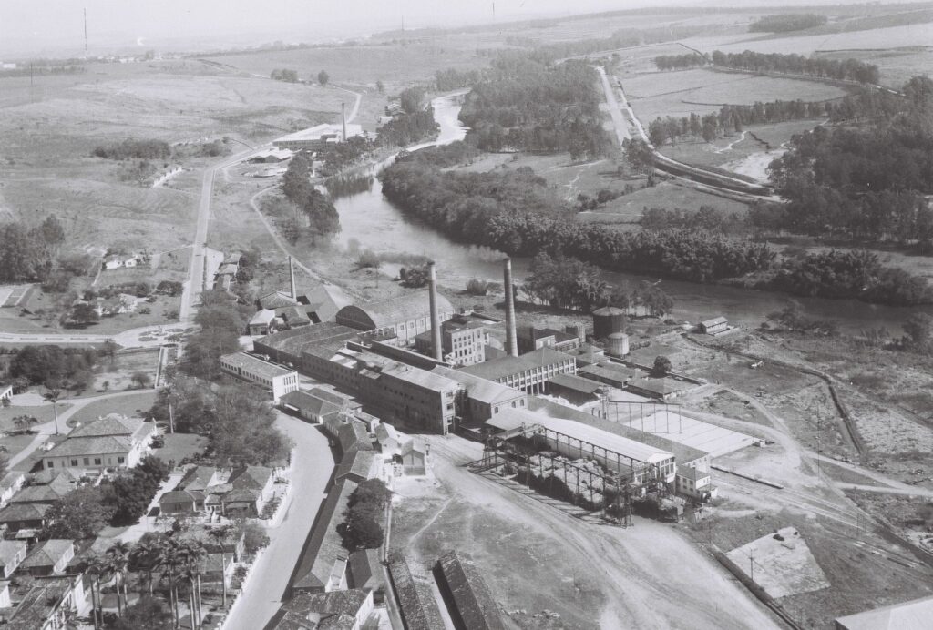 A fotografia mostra uma vista aérea em preto e branco de uma antiga fábrica ou usina industrial situada às margens de um rio. O complexo possui grandes galpões, duas chaminés altas e um reservatório cilíndrico, cercado por pequenas casas e ruas sinuosas. Ao fundo, vê-se uma paisagem rural ampla, com campos, árvores e estradas que se estendem até o horizonte.